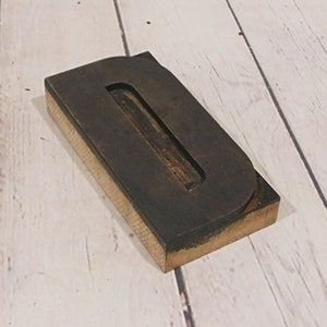 May include: A rectangular wooden letterpress block featuring a dark, raised letter O. The block has a natural light-colored wood base with a dark, weathered metal or composite top surface that forms the shape of the letter O in a bold, sans-serif font.