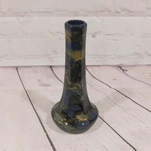 May include: A ceramic bud vase with a narrow neck and a flared base, featuring a glossy, marbled glaze in shades of deep navy blue, dark charcoal, and muted mustard yellow, creating a swirling, organic pattern across the entire surface.