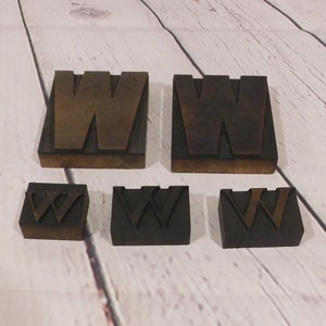 May include: A collection of five vintage metal letterpress blocks, each featuring the letter W in varying sizes. Two large blocks are positioned at the top, while three smaller blocks are arranged in a row below, all showing a dark, oxidized finish.