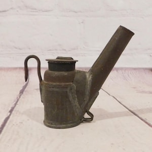 May include: Antique metal oil lamp with a handle and a long spout. The lamp has a dark, aged appearance, with visible signs of wear and tear. The design includes a curved handle and a small, circular lid. The lamp is likely made of iron or a similar metal.