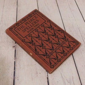 May include: A vintage hardcover book titled The Peirce Spellers Book Two by Ginn and Company. The cover is a muted reddish-brown color with black decorative filigree patterns and geometric lines. The title is printed in bold black capital letters.