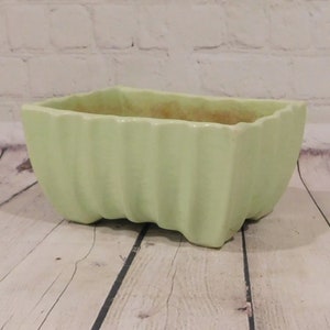 May include: A rectangular, light green ceramic planter with a ribbed exterior. The planter has a slightly textured interior and sits on four small feet. The background features a white brick wall and a wooden surface.