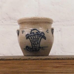 May include: A small, tan ceramic pot with a blue floral design. The pot has two small handles and a wide opening. The floral design is a basket with flowers. The pot is sitting on a wooden surface.