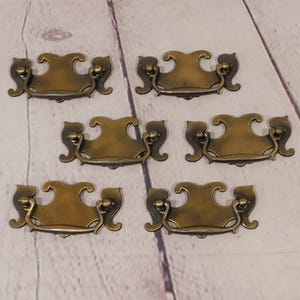 May include: A set of six brass-toned metal drawer pulls, each featuring a decorative backplate with curved, symmetrical edges and a hinged, swinging bail handle attached to two small circular mounting points on the sides of the plate.
