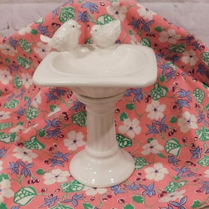 May include: A white ceramic birdbath featuring a square basin supported by a fluted pedestal base. Two small, detailed white ceramic birds are perched on the rim of the basin, facing each other. The entire structure is finished in a glossy white glaze.