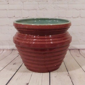 May include: A round ceramic planter with a glossy dark reddish-brown exterior featuring horizontal ribbed detailing. The interior of the pot is finished in a smooth, contrasting light teal glaze, with a wide, circular opening and a thick rim.