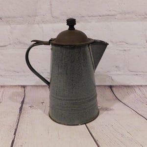 May include: A gray speckled enamel coffee pot with a dark brown lid and a small black knob on top. The pot features a curved handle on the side, a tapered body with horizontal ridges near the base, and a triangular spout for pouring liquids.