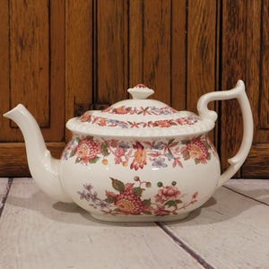 May include: A cream-colored ceramic teapot featuring a colorful floral pattern with pink, red, and blue blossoms. The teapot has a curved handle, a spout, and a matching lid with a small knob. The floral design wraps around the body and lid.
