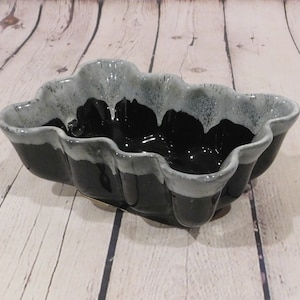 May include: A ceramic, scalloped-edge bowl with a glossy black interior and a gray and black exterior. The bowl has a unique, wavy rim and sits on a wooden surface. This decorative bowl is ideal for serving or display.