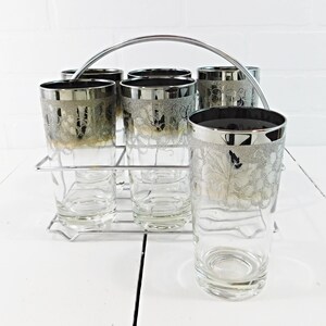 May include: A set of six clear glass tumblers with silver rims and a silver leaf pattern around the upper portion, held in a chrome wire carrier with a curved handle. Each glass features a metallic silver band at the top and a textured leaf design.