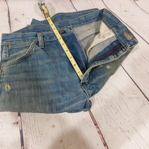 May include: Light blue denim jeans with a measuring tape showing a 4-inch measurement. The jeans have a classic five-pocket design, a zipper fly, and distressed details. The jeans are folded on a light wood surface.