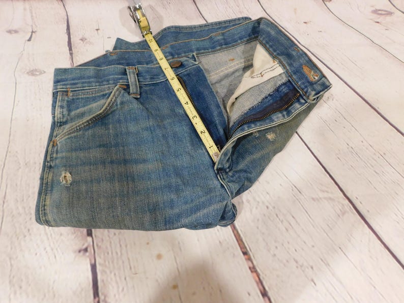 May include: Light blue denim jeans with a measuring tape showing a 4-inch measurement. The jeans have a classic five-pocket design, a zipper fly, and distressed details. The jeans are folded on a light wood surface.