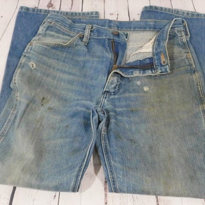May include: Faded blue denim jeans with a zipper fly and button closure. The jeans show signs of wear, including discoloration and small holes. The jeans are laid flat on a light-colored wooden surface.