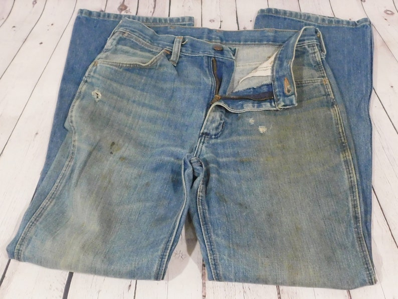 May include: Faded blue denim jeans with a zipper fly and button closure. The jeans show signs of wear, including discoloration and small holes. The jeans are laid flat on a light-colored wooden surface.