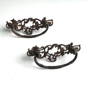 May include: Two matching drawer pulls made of dark, aged bronze-toned metal. Each pull features an intricate, ornate filigree backplate with scrolling patterns and a curved, textured drop handle that hangs from two circular mounting points.