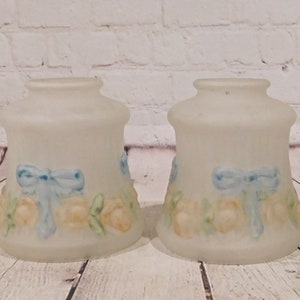 May include: Two frosted glass lamp shades. Each shade features a painted design of yellow roses, green leaves, and a blue bow. The shades have a bell-like shape and are suitable for a vintage or cottage-style decor.