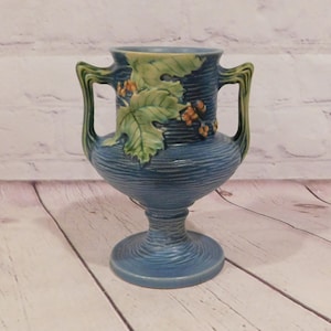 May include: Blue ceramic vase with two handles and a pedestal base. The vase features a textured surface and is decorated with green leaves and orange berries. The handles and the rim of the vase are green.