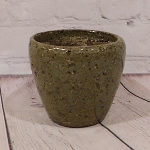 May include: A speckled, olive-green ceramic planter with a rounded shape. The planter has a slightly textured surface and a wide opening. The interior appears to contain soil or other planting material. The background features a white brick wall and a wooden surface.