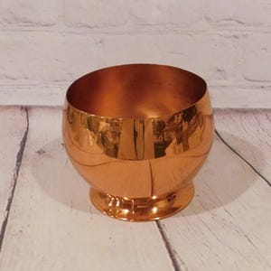 May include: A polished copper bowl with a rounded, spherical body and a circular pedestal base. The metallic surface features a warm, reflective orange-brown tone with visible highlights and a smooth, seamless finish throughout the entire object.
