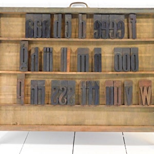May include: A rectangular wooden letterpress type case featuring three horizontal shelves. The case contains various dark, weathered wooden letter blocks arranged in rows. The frame is constructed from light brown wood with a metal handle on top.