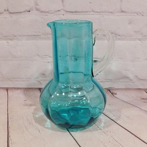 May include: A translucent turquoise glass pitcher featuring a bulbous base with a textured, paneled design and a smooth, clear glass handle. The vessel has a wide, cylindrical neck and a small pouring spout at the top rim, crafted from glass.