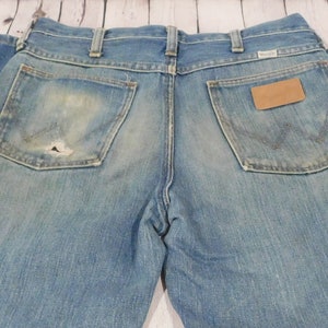 May include: Light blue denim jeans with a worn appearance, featuring two back pockets and a small leather patch. The jeans show signs of wear and tear, including a hole in one of the pockets. The Wrangler logo is visible.