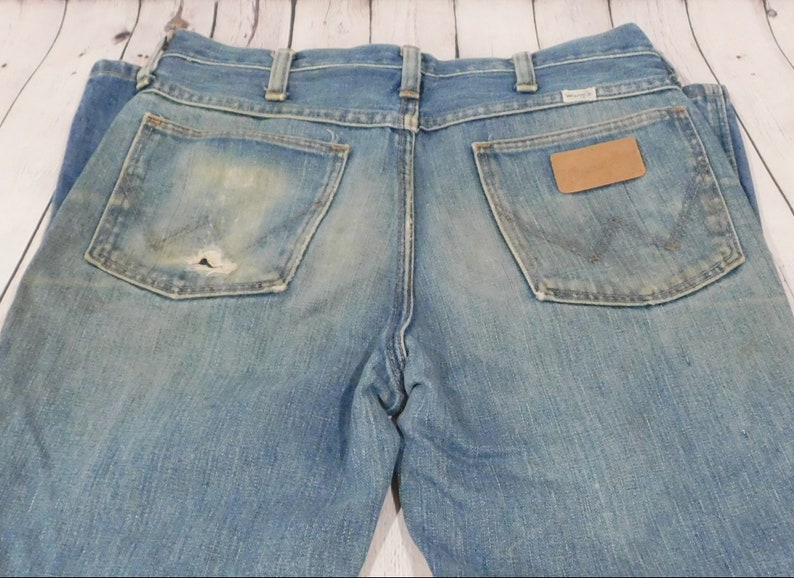May include: Light blue denim jeans with a worn appearance, featuring two back pockets and a small leather patch. The jeans show signs of wear and tear, including a hole in one of the pockets. The Wrangler logo is visible.