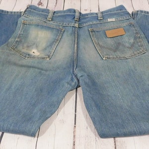 May include: Light blue denim jeans with a worn appearance. The jeans feature a classic five-pocket design, a brown leather patch, and the Wrangler logo. The back pocket has a distinctive stitching pattern. The jeans are folded.
