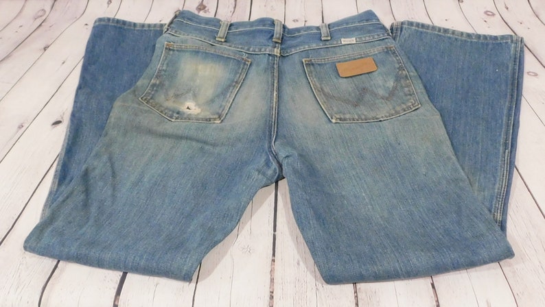 May include: Light blue denim jeans with a worn appearance. The jeans feature a classic five-pocket design, a brown leather patch, and the Wrangler logo. The back pocket has a distinctive stitching pattern. The jeans are folded.