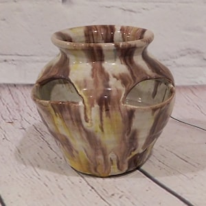 May include: A ceramic vase with a unique design. The vase features a marbled pattern of brown, yellow, and cream colors. It has two openings on the sides, and a wide opening at the top. The vase is likely handmade.