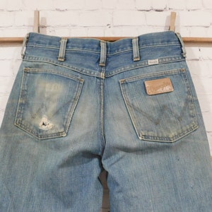 May include: Light blue denim Wrangler jeans with a distressed look. The jeans have a worn patch on the back pocket and a leather Wrangler patch. The jeans are hanging on a wooden rod with a white brick background.
