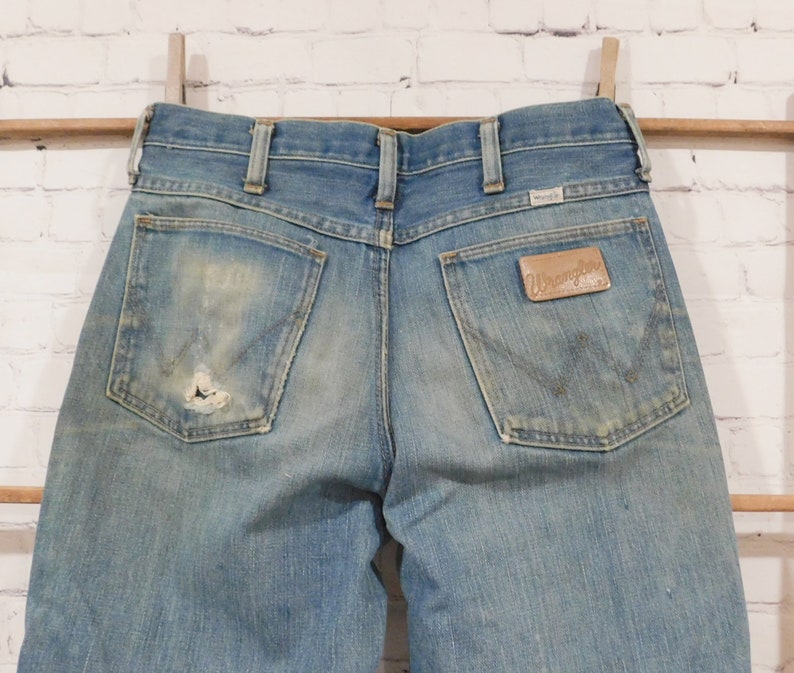 May include: Light blue denim Wrangler jeans with a distressed look. The jeans have a worn patch on the back pocket and a leather Wrangler patch. The jeans are hanging on a wooden rod with a white brick background.