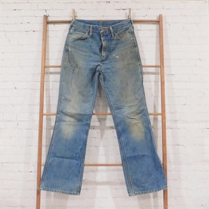 May include: Light blue denim jeans with a faded wash and slight distressing. The jeans are hanging on a wooden ladder against a white brick wall. The jeans have a classic five-pocket design and a flared leg.