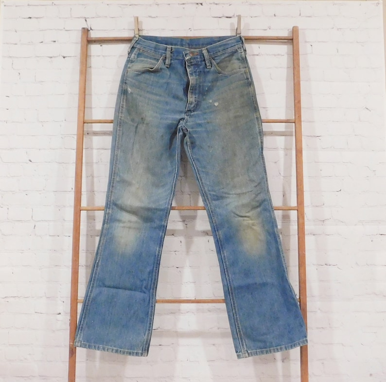 May include: Light blue denim jeans with a faded wash and slight distressing. The jeans are hanging on a wooden ladder against a white brick wall. The jeans have a classic five-pocket design and a flared leg.