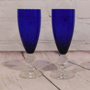 May include: A pair of cobalt blue glass champagne flutes featuring a clear, textured, spherical glass accent on the stem and a circular clear glass base. The bowls are deep blue, while the stems and bases are made of transparent, colorless glass.