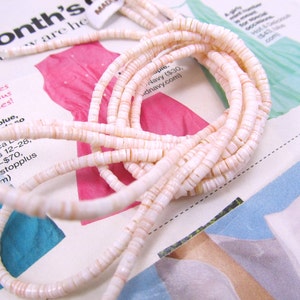 May include: A strand of white and pink shell beads, arranged in a circular pattern on a magazine page. The beads are small and uniform in size, and they have a slightly iridescent sheen.