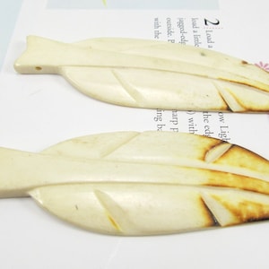 May include: Two cream-coloured, leaf-shaped pendants with carved details and a small hole for stringing. The edges have a subtle brown gradient. The pendants are lying on a white surface with text.