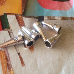 May include: Six silver metal cone-shaped cord ends with a decorative design. The cord ends are arranged in a cluster on a light brown surface.