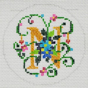 May include: Cross-stitch embroidery of the letter N with a floral design. The letter is gold, the flowers are blue, pink, yellow, and green, and the vines are green.