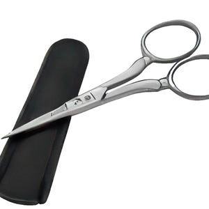 May include: A pair of small, silver-colored scissors with rounded finger holes, resting next to a black, rectangular sheath. The scissors have a curved design and pointed blades, suitable for detailed cutting tasks.
