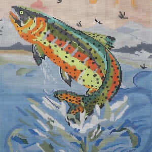 May include: A needlepoint image of a jumping trout. The fish has orange, green, and yellow markings. The background features a light blue water scene, a peach sky, and small black birds.