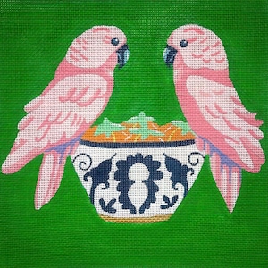 May include: A canvas painting featuring two pink parrots facing each other above a decorative bowl. The bowl has a white base with blue floral designs and contains orange and green elements. The background is a solid green.