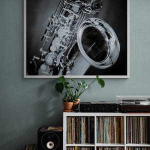 May include: Black and white framed print of a saxophone. The saxophone is silver and highly detailed, with a large bell and intricate keys. The print is displayed above a white shelf filled with vinyl records.