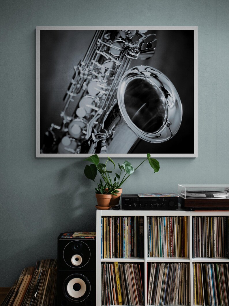 May include: Black and white framed print of a saxophone. The saxophone is silver and highly detailed, with a large bell and intricate keys. The print is displayed above a white shelf filled with vinyl records.