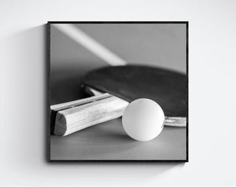 May include: A black and white photograph featuring a table tennis paddle with a wooden handle and a smooth, dark rubber surface, positioned next to a round, white table tennis ball. The composition is square and framed in a thin black border.