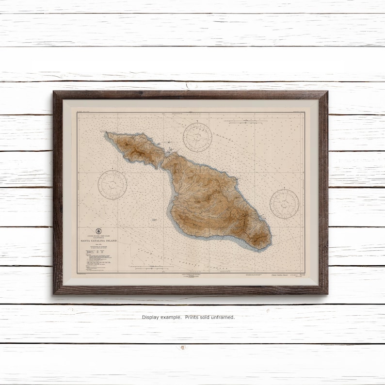 May include: A framed nautical chart of Santa Catalina Island, California, featuring detailed topographical brown shading for the landmass, blue depth soundings, and three compass roses. The map includes printed text and technical data.