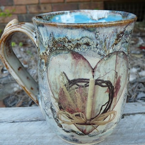 May include: A ceramic mug with a rustic, distressed finish. The mug features a heart-shaped design with a floral motif and a brown ribbon. The mug has a light blue glaze and a brown handle.