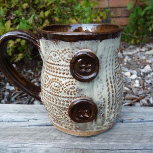 May include: A ceramic mug with a glossy dark brown handle and rim, featuring a cream-colored body with a textured knit-like pattern. Two dark brown, circular, four-hole buttons are attached to the side of the mug, integrated into the surface.