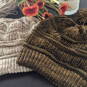 May include: Two handmade knitted beanies in neutral tones. One is a light beige, the other a dark brown with a textured stitch pattern. Knitting needles and yarn are visible, suggesting a craft theme. The beanies are likely for cold weather.
