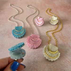 May include: A collection of shell-shaped lockets and rings in blue, pink, and white. The lockets hang on silver or gold chains, while the rings feature a hinged design that opens to reveal a small compartment. Each piece has a scalloped texture.